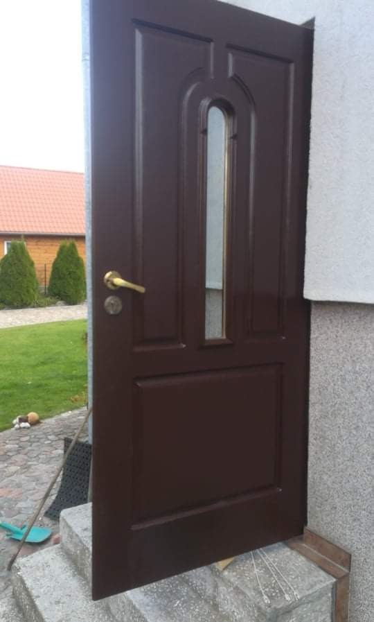 Brązowe drzwi wejściowe z szybką, zamontowane na zewnątrz budynku, widoczne schody wejściowe i fragment trawnika z drzewami.