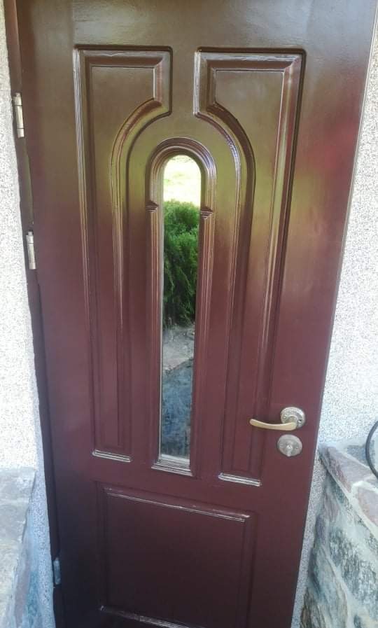 Ciemnobrązowe drzwi wejściowe z ozdobnym przeszkleniem, widok na zieleń ogrodu przez szybę, srebrna klamka i zamek, fragment ściany z szarego kamienia po prawej stronie.