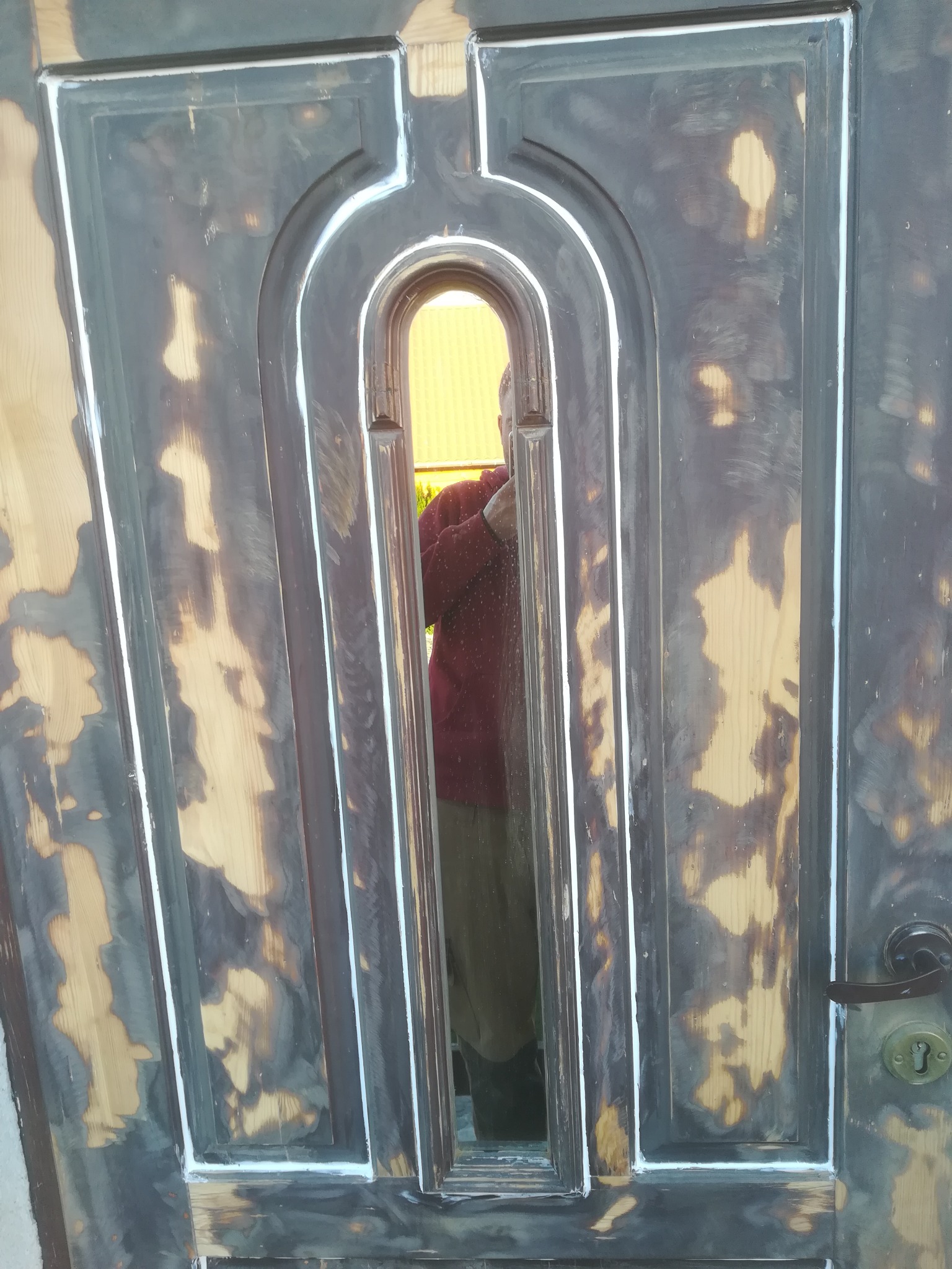 Drzwi drewniane z szybą po częściowym usunięciu starej farby, z widocznymi śladami szpachli i białego silikonu na krawędziach, z odblaskiem osoby w szybie.