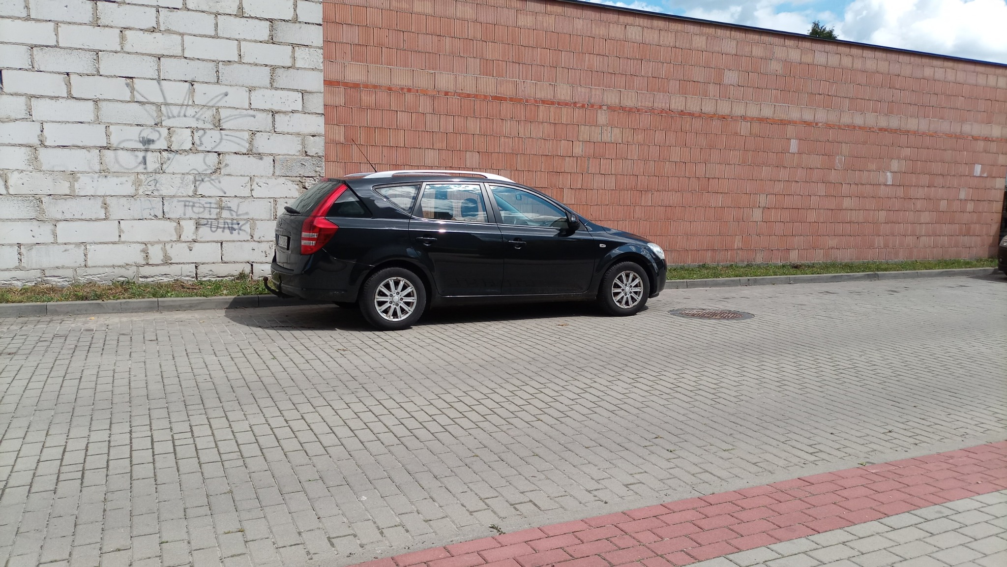 Czarny samochód kombi Kia Ceed zaparkowany na kostce brukowej przed dwiema ścianami: białą z graffiti i czerwoną z cegły kratówki.