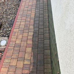 ,,GLANZARO''JAKUB WOJTÓŚCISZYN - Częściowo wyczyszczona kostka brukowa w odcieniach czerwieni i brązu, kontrastująca z brudną częścią. Widoczne kamienie i fragment ściany budynku.