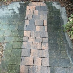 ,,GLANZARO''JAKUB WOJTÓŚCISZYN - Częściowo wyczyszczona kostka brukowa, widoczna różnica między brudną, zieloną powierzchnią a czystą, mokrą kostką. W rogu kadru widoczny fragment buta.