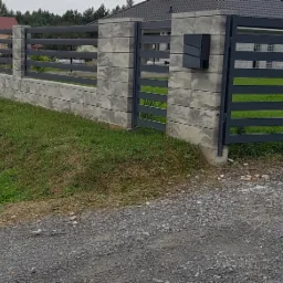 Nowoczesne ogrodzenie panelowe z betonowymi słupkami i skrzynką na listy, widok z poziomu gruntu na tle zieleni i domu.