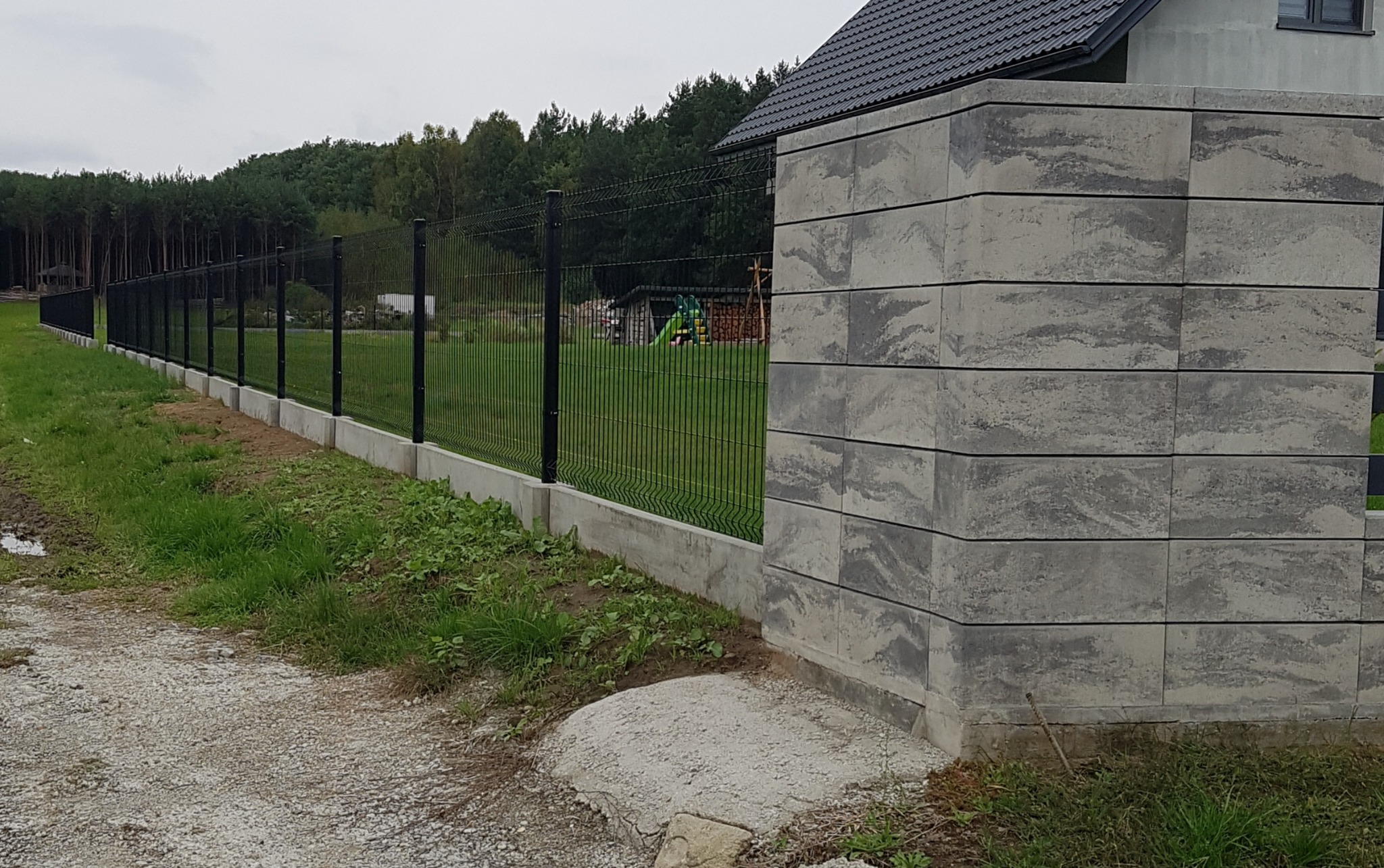 Nowoczesne ogrodzenie panelowe z podmurówką betonową i słupkiem z szarego betonu architektonicznego, widok z zewnątrz posesji na tle lasu i domu.