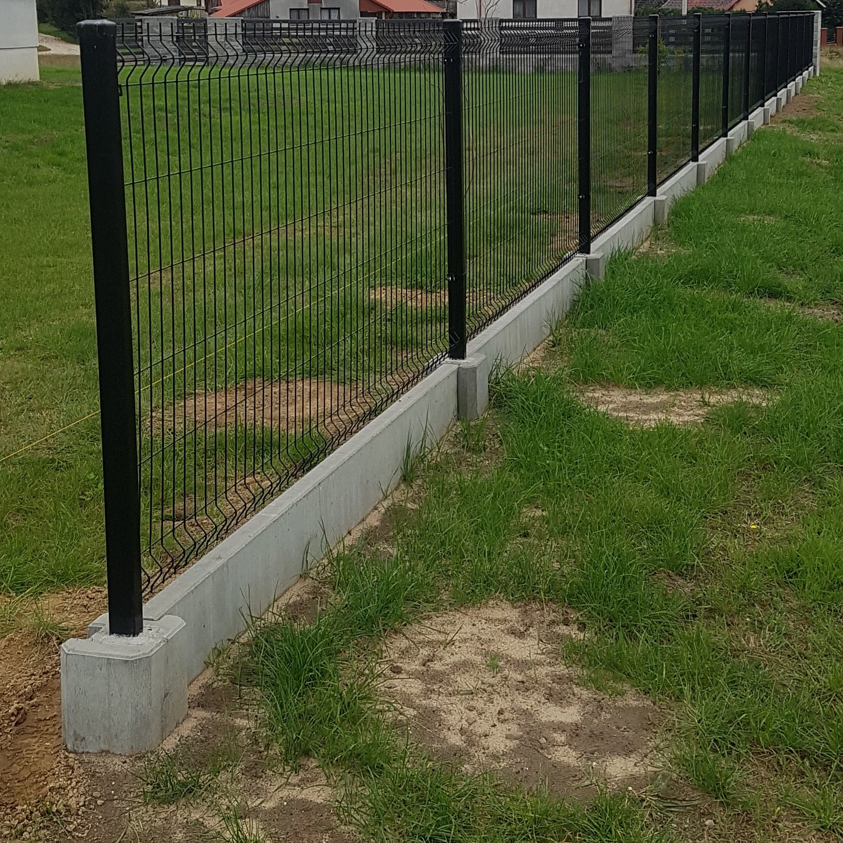 Nowo zainstalowane, czarne, panelowe ogrodzenie z podmurówką betonową na trawiastym terenie.