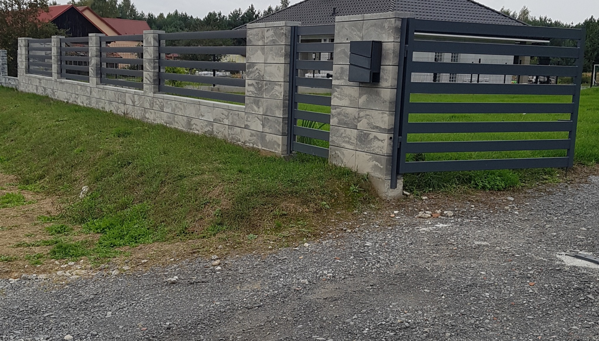 Nowoczesne ogrodzenie panelowe z betonowymi słupkami i skrzynką na listy, widok z poziomu gruntu na tle zieleni i domu.