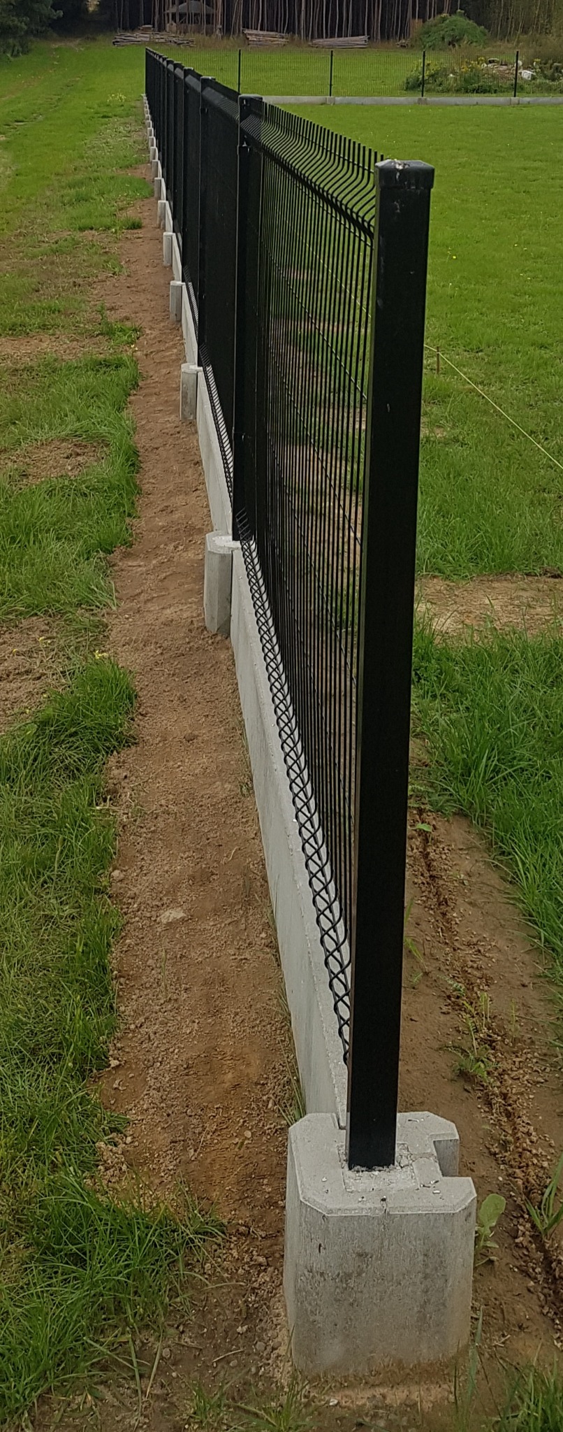 Długi, czarny, metalowy płot panelowy z podmurówką betonową, ustawiony na trawiastym terenie, widok z boku, z perspektywą zbieżną.