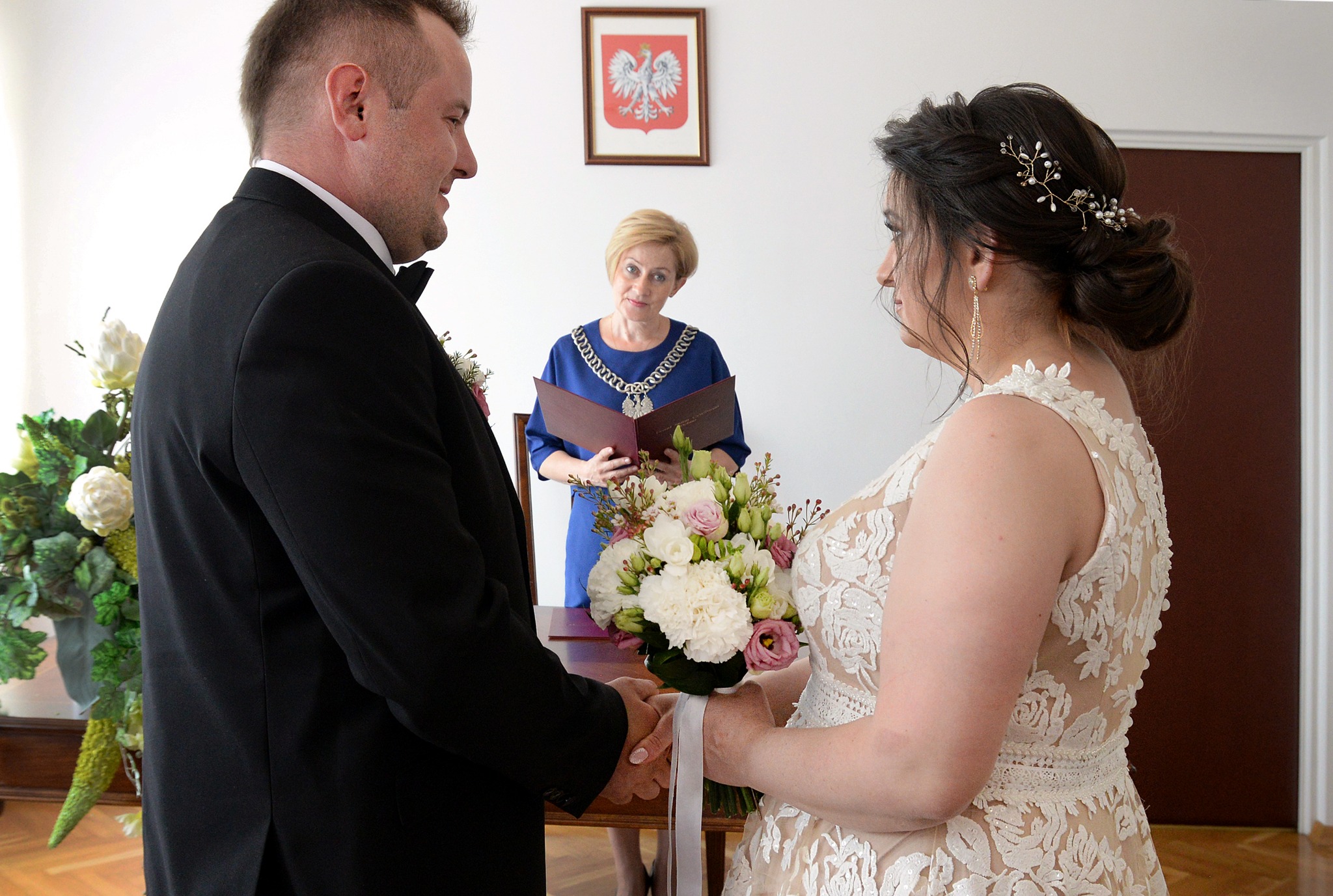 Para młoda trzymająca się za ręce podczas ceremonii ślubnej w urzędzie stanu cywilnego, urzędniczka w tle, bukiet ślubny z białych i różowych kwiatów.