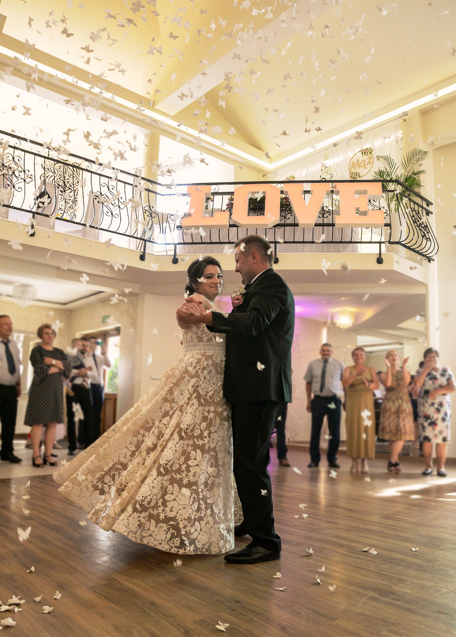 Para młoda tańczy podczas wesela, spadają białe motyle, w tle goście i dekoracja z napisem LOVE.