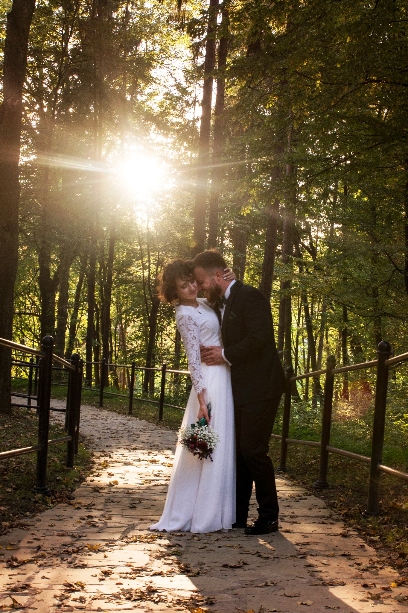 Para młoda obejmująca się na leśnej ścieżce, panna młoda w sukni z koronkowymi rękawami trzyma bukiet, promienie słońca przebijają przez drzewa.