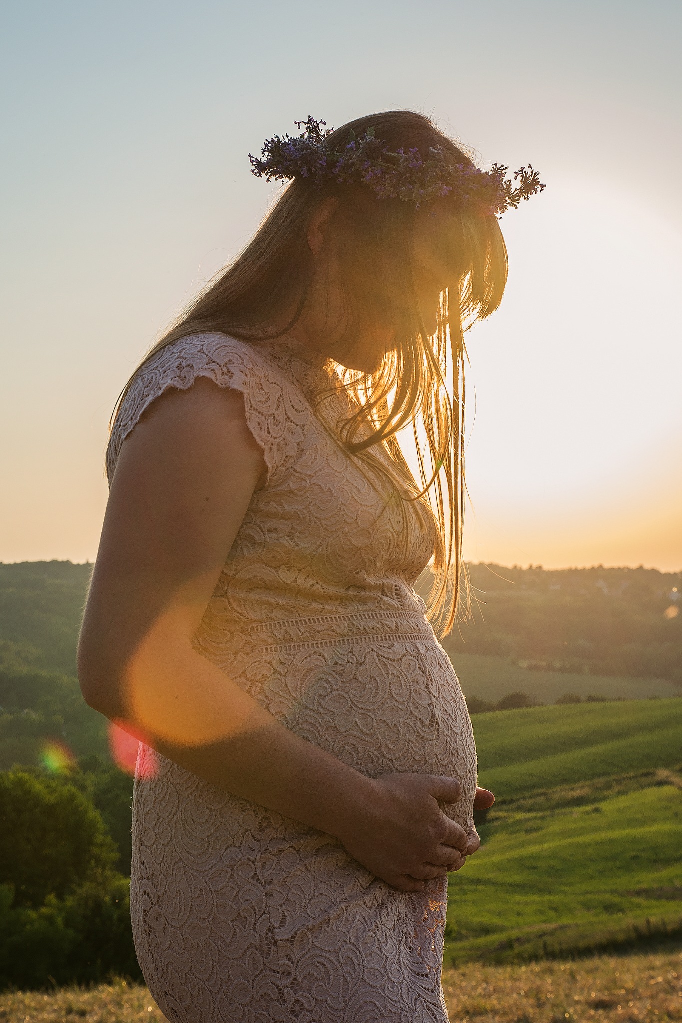 Kobieta w ciąży w koronkowej sukience i wianku z polnych kwiatów, delikatnie obejmująca brzuch na tle zachodzącego słońca i krajobrazu Brzeska.