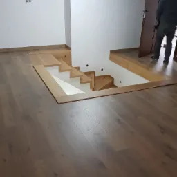 Montaż drewnianej antresoli z widocznymi schodami i wykończeniem podłogi, kabel zwisający z sufitu, częściowo widoczna osoba w tle.