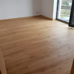 Jasna, drewniana podłoga ułożona w pustym pokoju z białymi ścianami i dużym oknem balkonowym. Widoczne listwy przypodłogowe w kolorze podłogi.