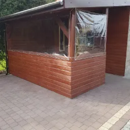 Drewniana konstrukcja z przezroczystymi zasłonami na zewnątrz domu, widok z boku na brukowaną powierzchnię.