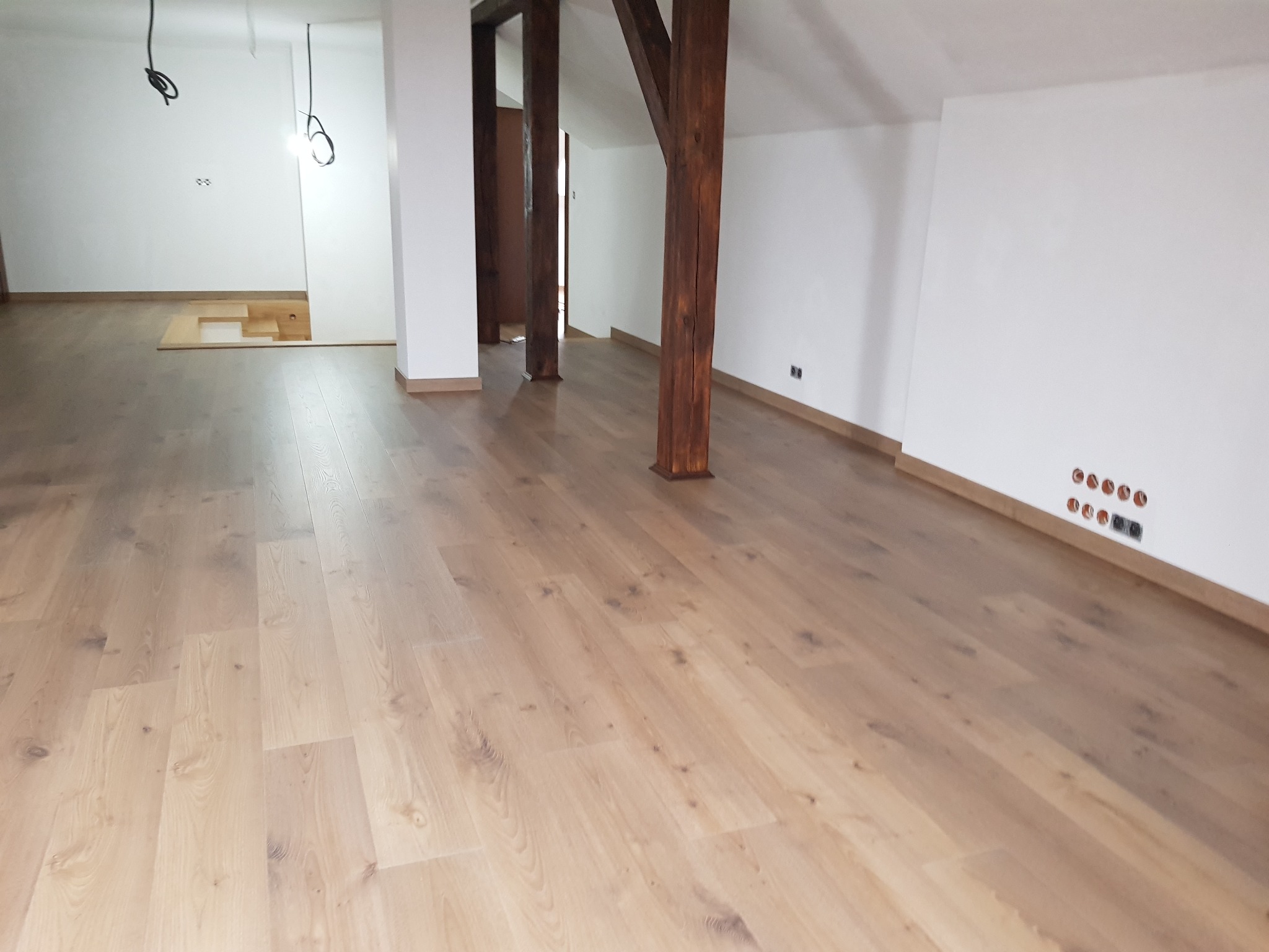 Jasne, drewniane deski podłogowe ułożone w przestronnym pokoju na poddaszu z drewnianymi belkami konstrukcyjnymi i białymi ścianami, widoczne gniazdka elektryczne w ścianie.