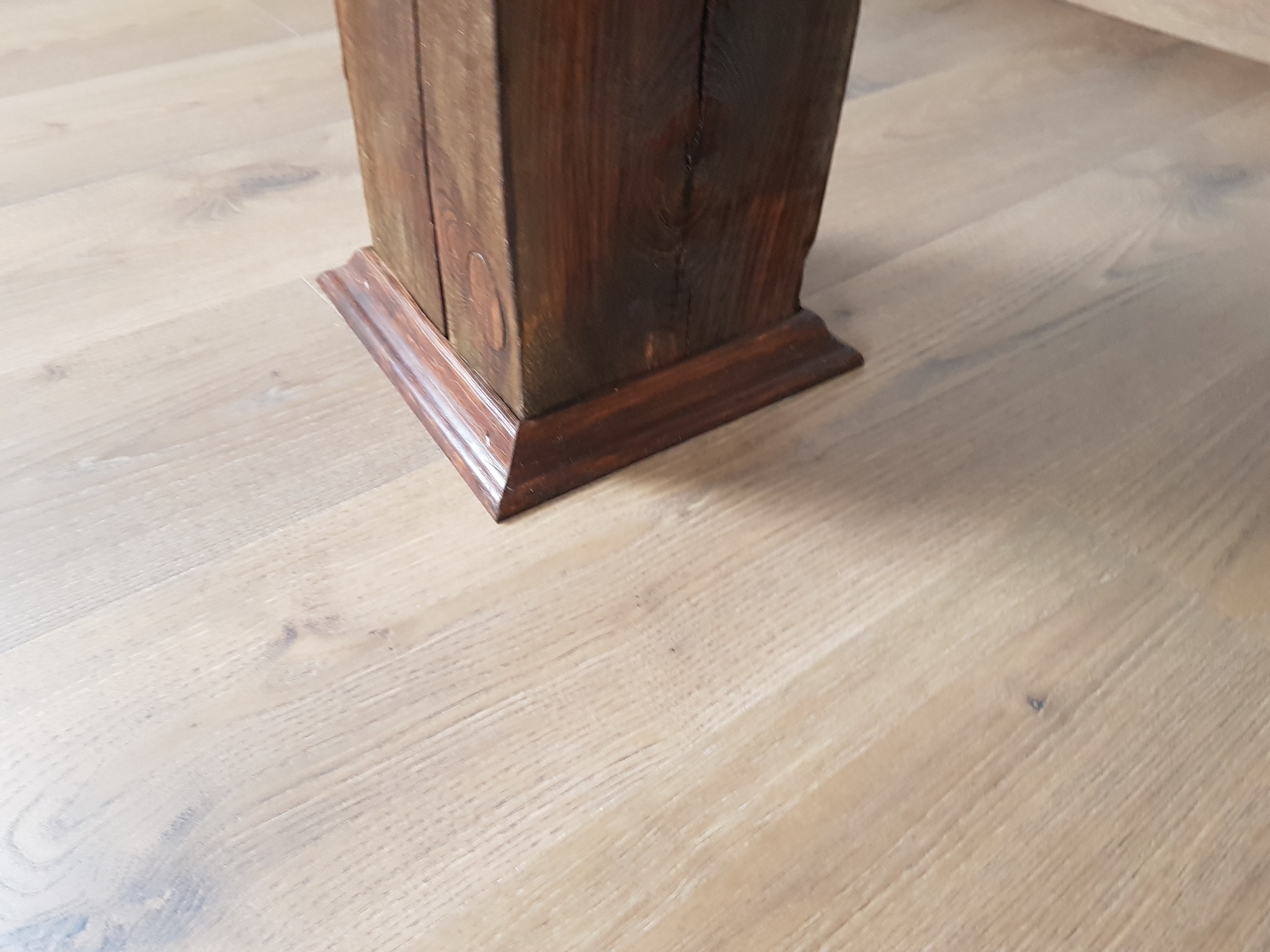 Fragment drewnianej kolumny z ciemnego drewna na jasnej, drewnianej podłodze z widocznymi słojami.