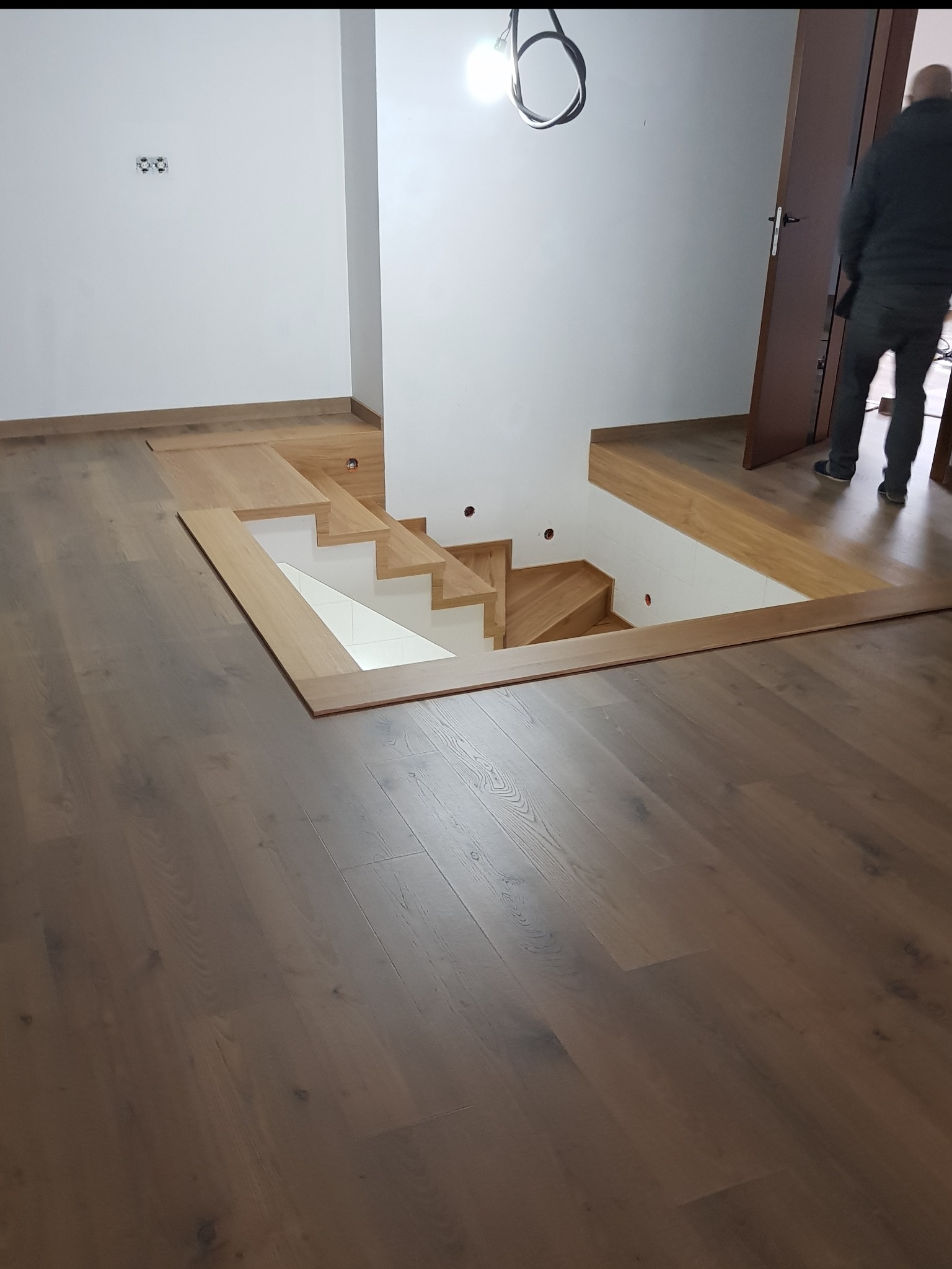 Montaż drewnianej antresoli z widocznymi schodami i wykończeniem podłogi, kabel zwisający z sufitu, częściowo widoczna osoba w tle.