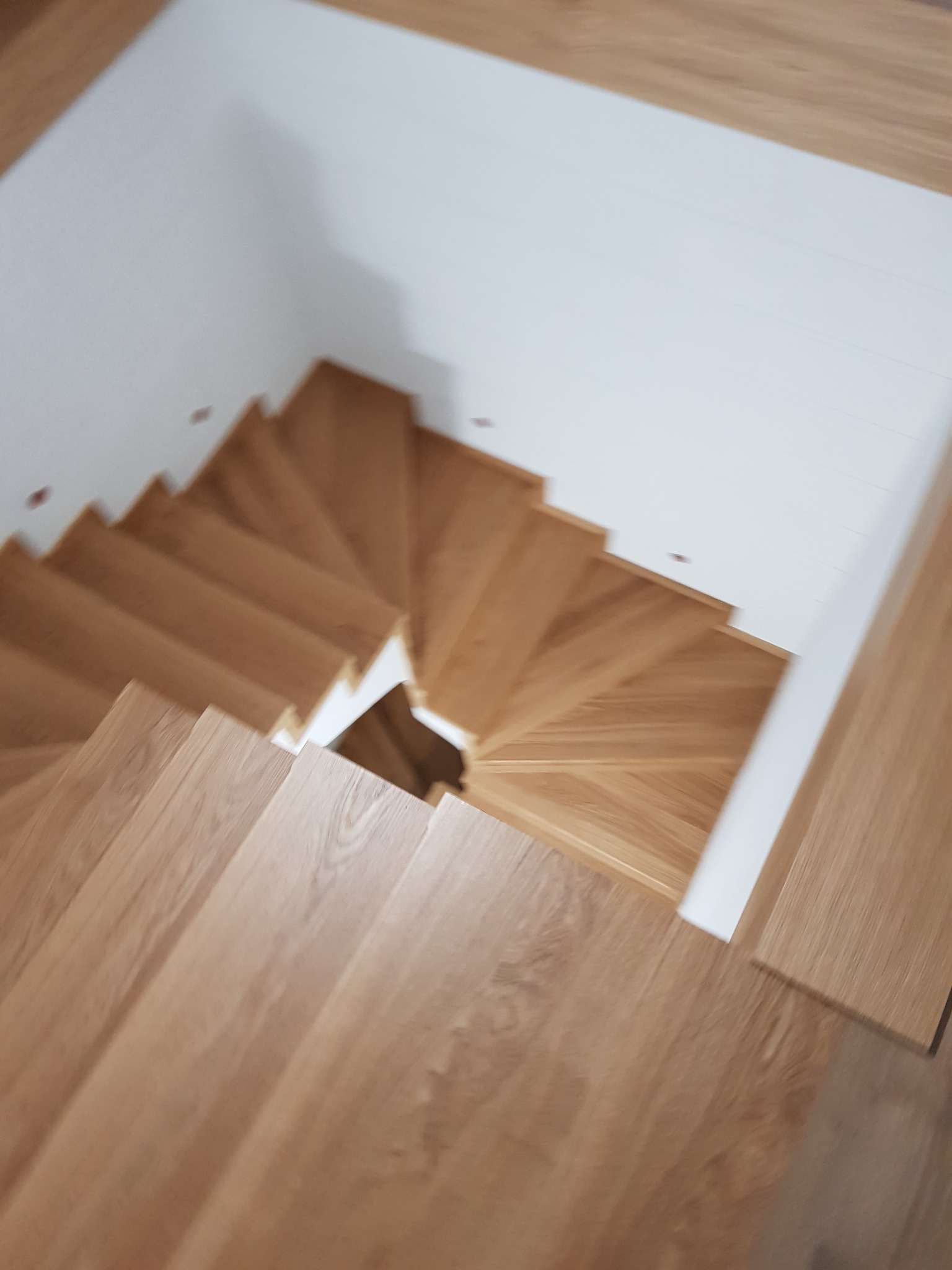 Widok z góry na drewniane schody spiralne z dębowymi stopniami, prowadzące w dół, z białymi ścianami w tle.