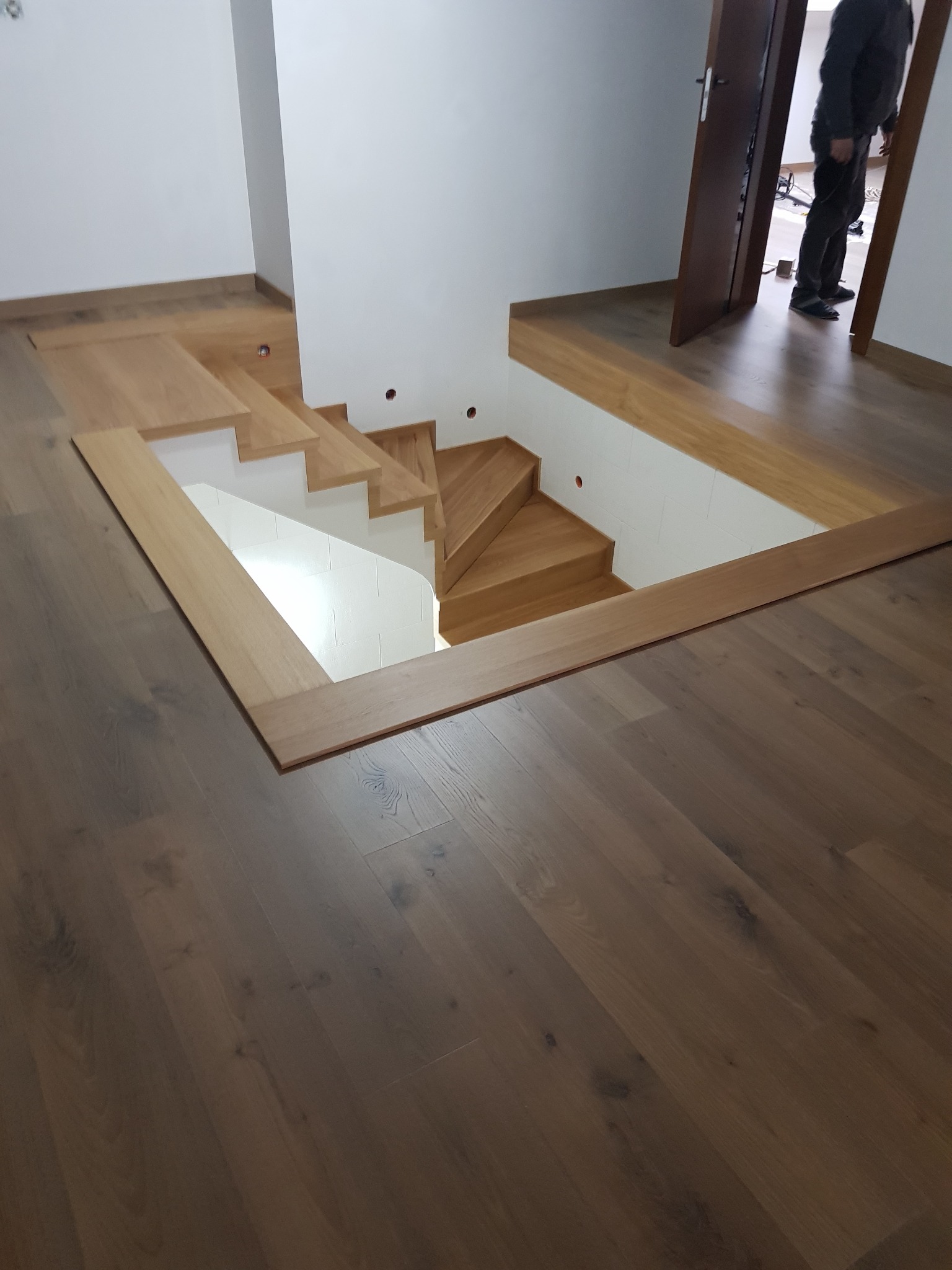 Nowoczesne drewniane schody prowadzące w dół, otoczone ramą z jasnego drewna, wkomponowane w ciemną drewnianą podłogę, widok z góry, w tle fragment otwartych drzwi z sylwetką osoby.