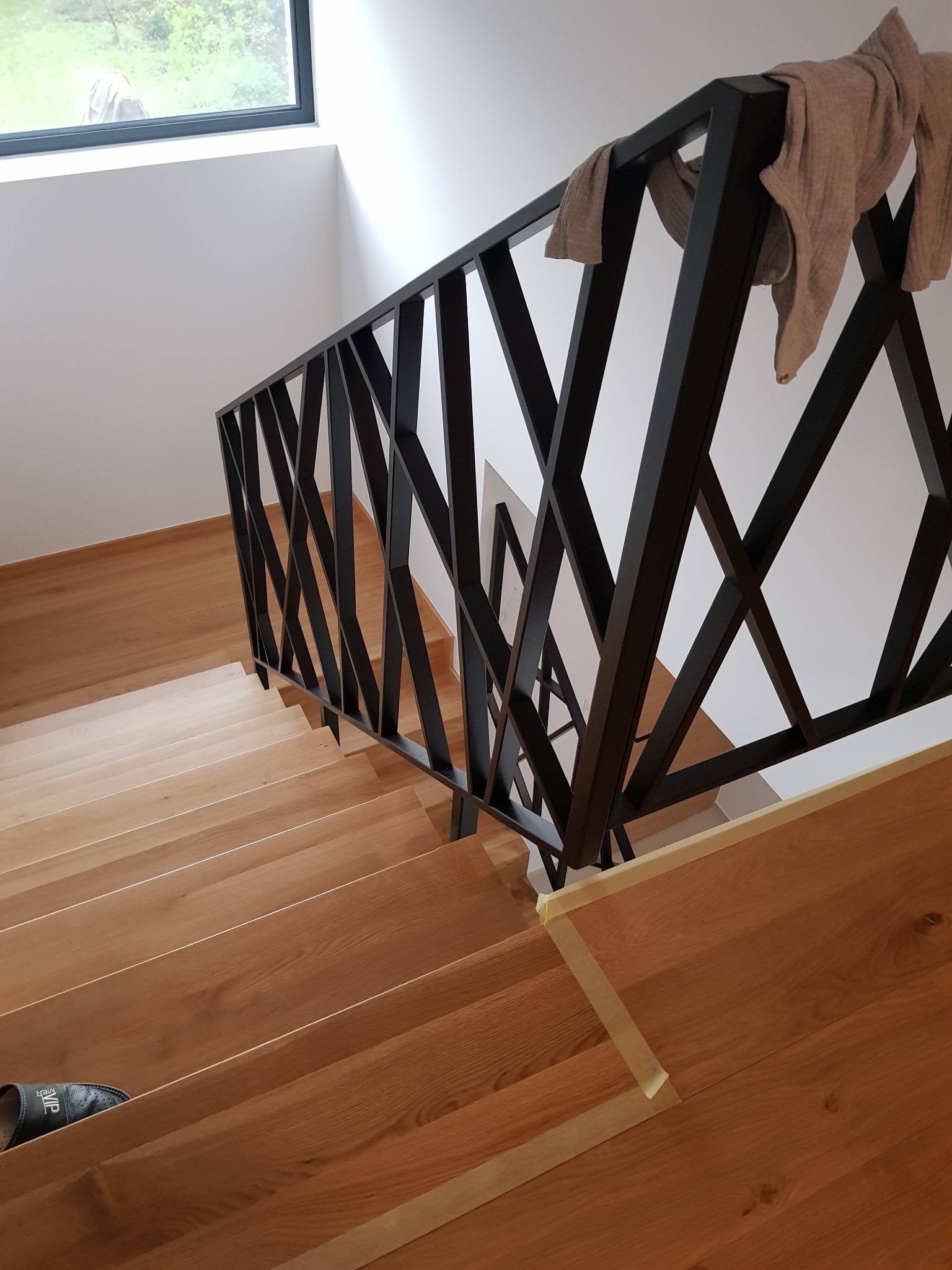 Schody drewniane z nowoczesną metalową balustradą o geometrycznym wzorze, zabezpieczone taśmą malarską na górnym stopniu, z szarą bluzą wiszącą na balustradzie i pojedynczym kapciem na dolnych...