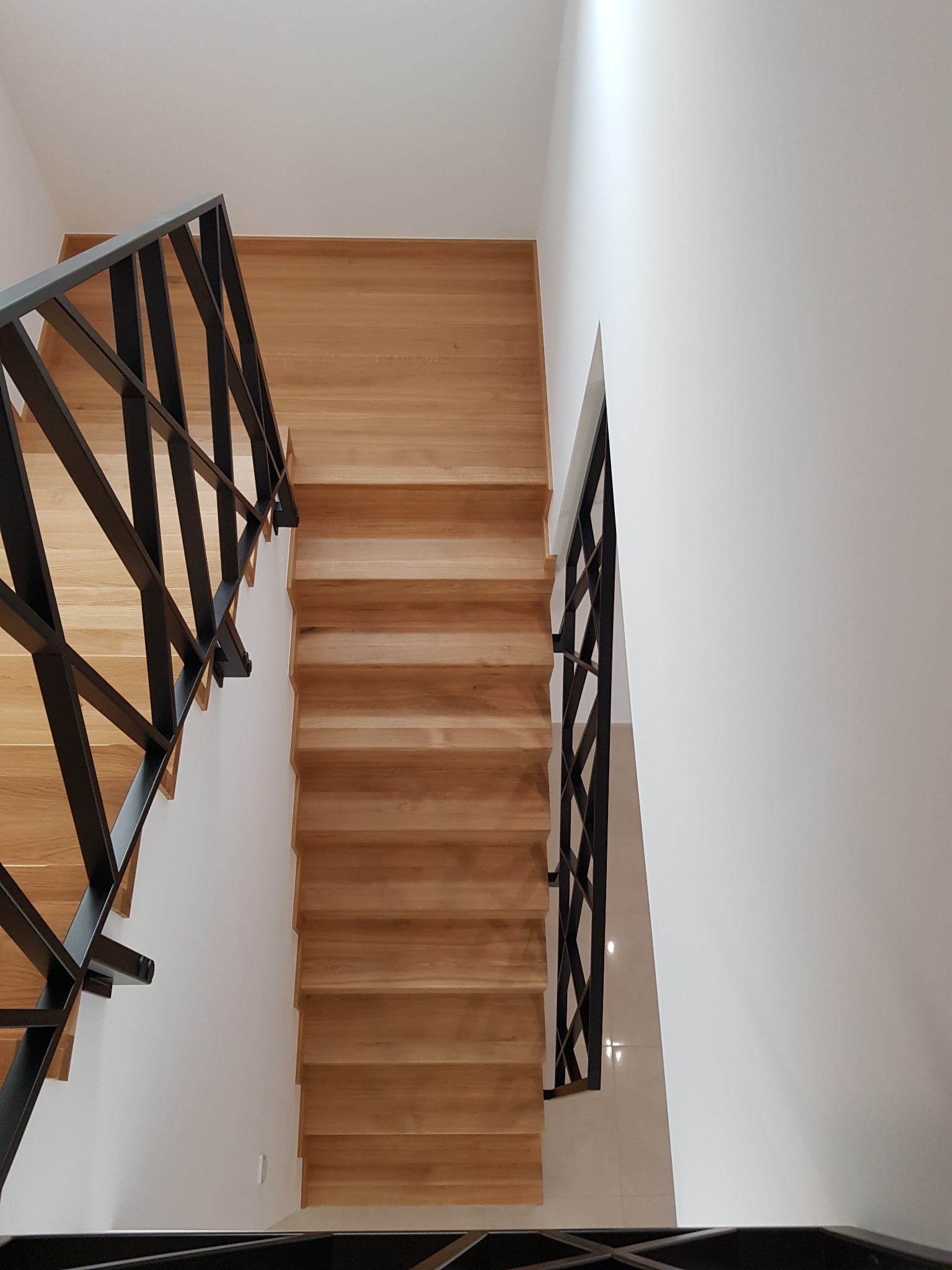 Widok z góry na drewniane schody z czarną, metalową balustradą o geometrycznym wzorze, prowadzące na białą, błyszczącą podłogę.