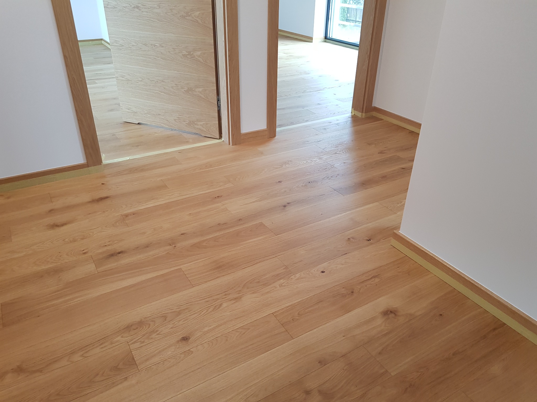 Jasna, drewniana podłoga ułożona w nowym domu, widoczne otwarte drzwi z drewnianymi ościeżnicami i listwy przypodłogowe zabezpieczone taśmą malarską.