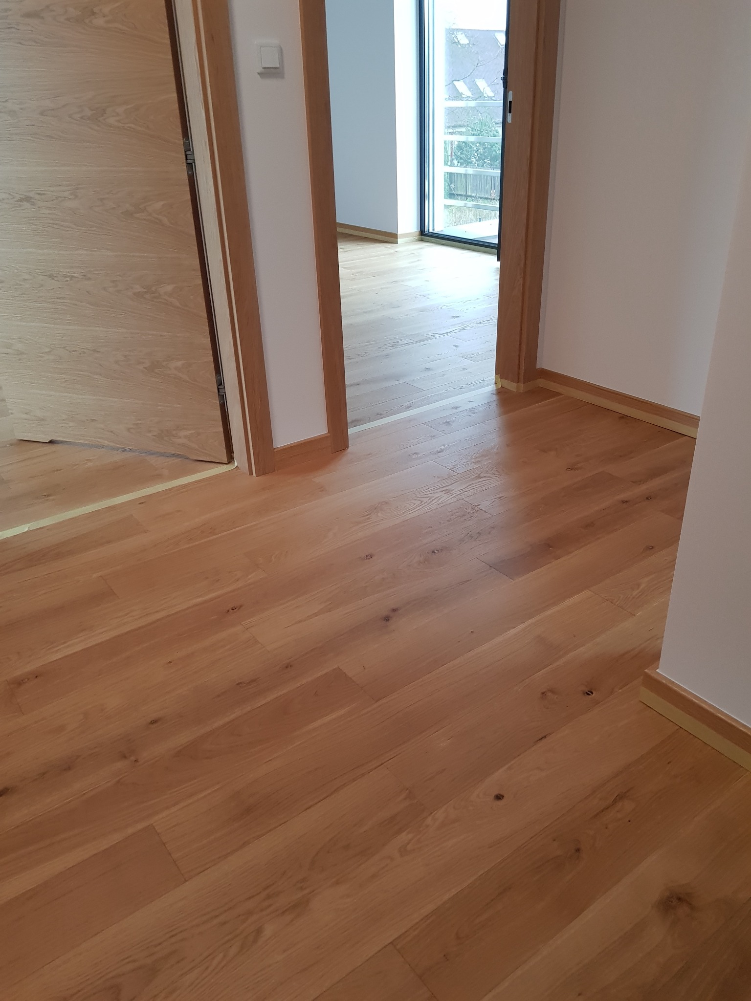 Jasne, dębowe panele podłogowe ułożone w korytarzu, widoczne drzwi z drewnianą ościeżnicą i fragment listwy przypodłogowej zabezpieczonej taśmą malarską.
