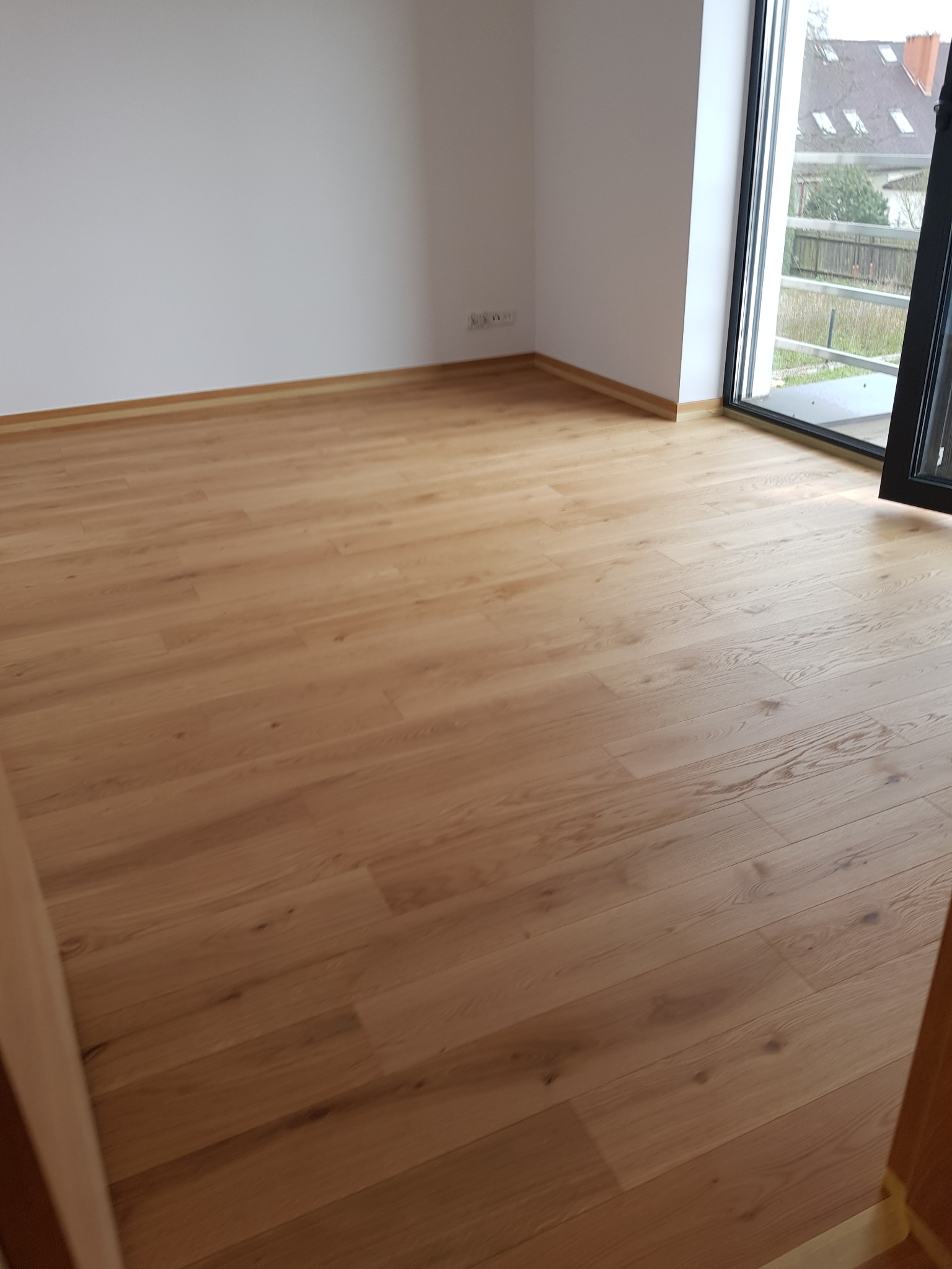 Jasna, drewniana podłoga ułożona w pustym pokoju z białymi ścianami i dużym oknem balkonowym. Widoczne listwy przypodłogowe w kolorze podłogi.
