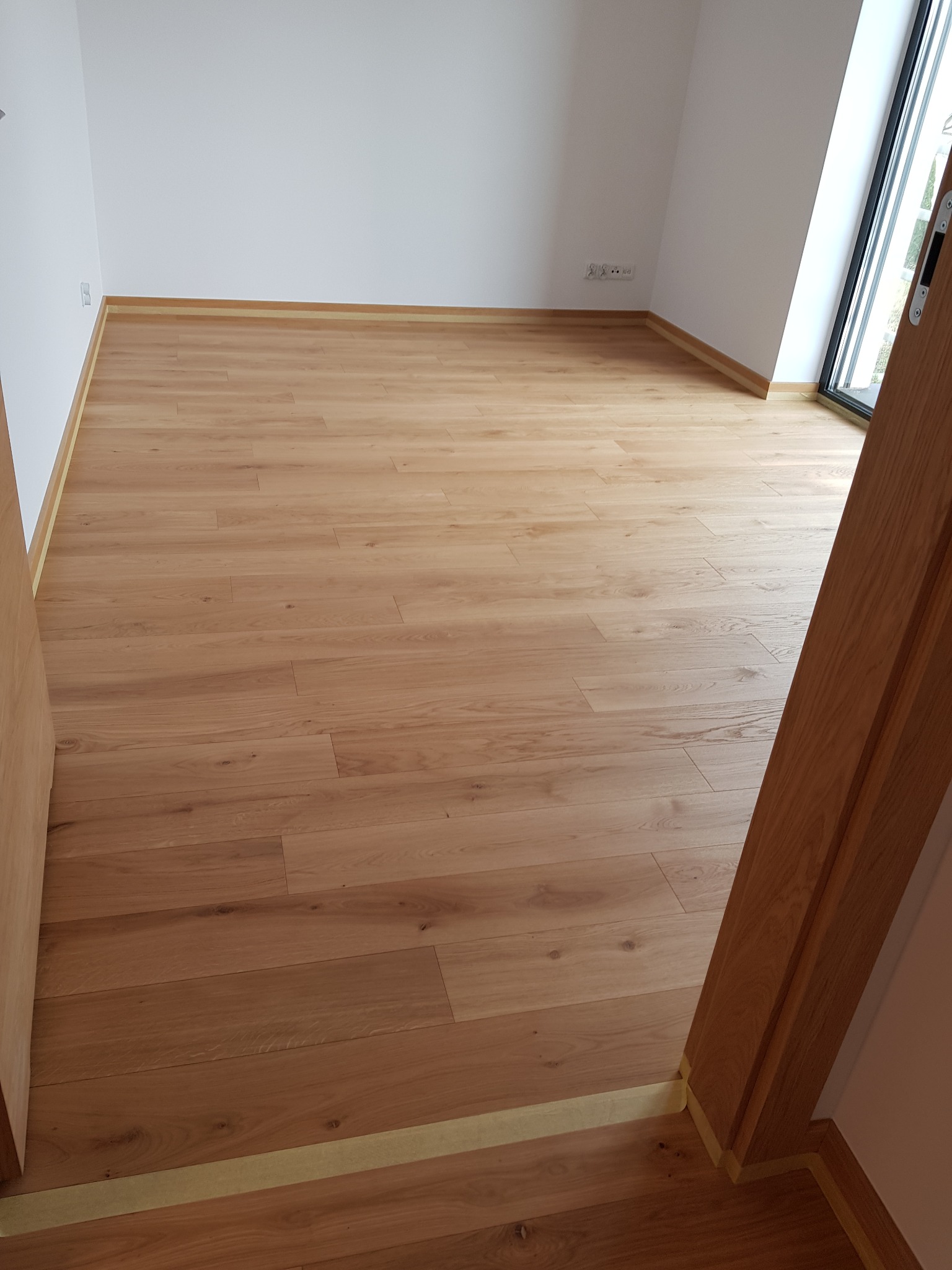 Jasne drewniane deski podłogowe ułożone w pustym pokoju, widoczna listwa przypodłogowa i fragment ościeżnicy z taśmą malarską zabezpieczającą podczas prac wykończeniowych.
