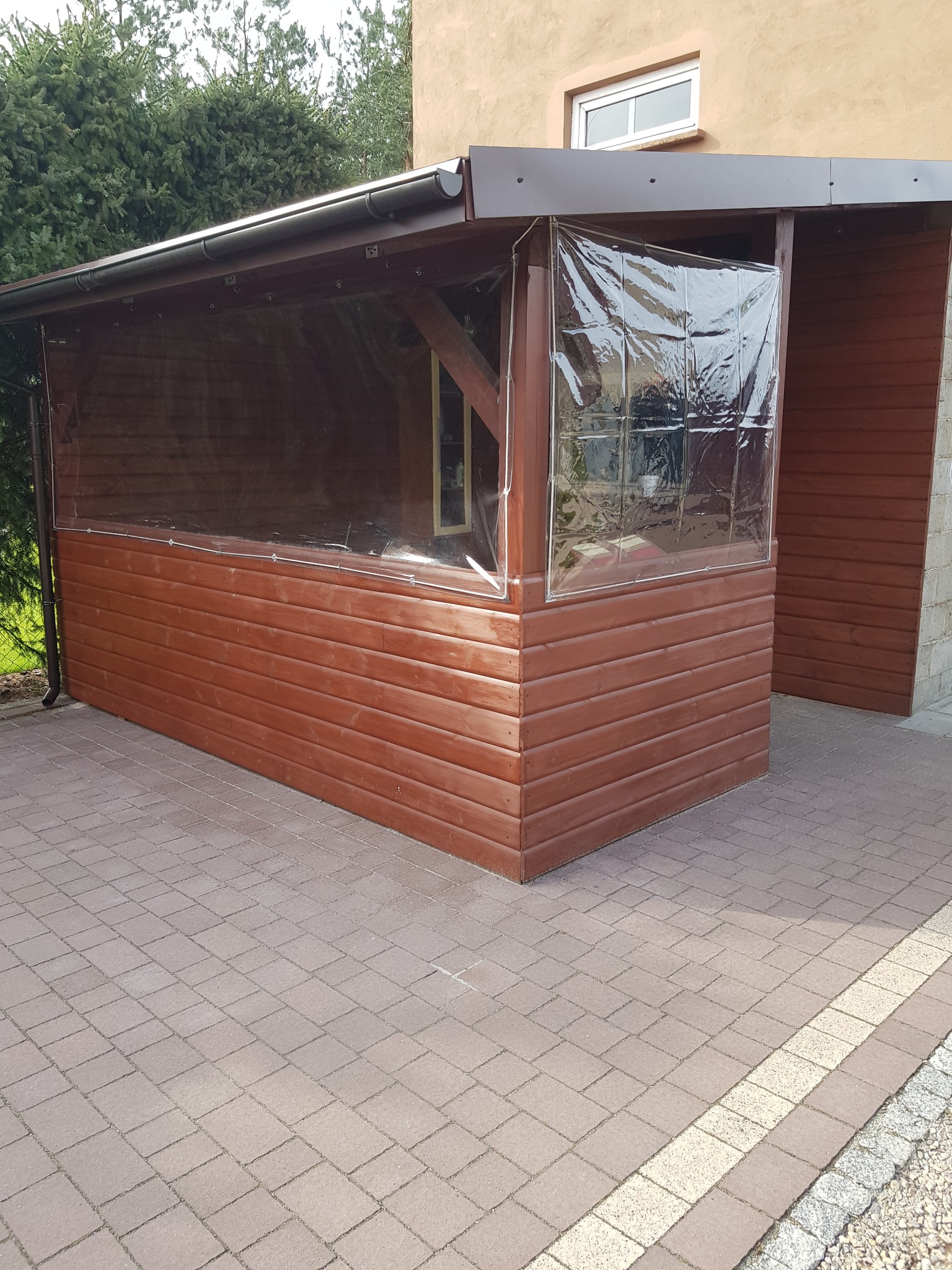 Drewniana konstrukcja z przezroczystymi zasłonami na zewnątrz domu, widok z boku na brukowaną powierzchnię.