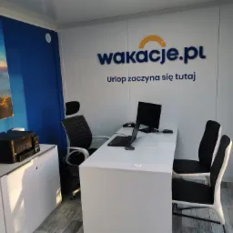 Wnętrze biura z logo 'wakacje.pl' na ścianie, biurko z laptopem i monitorem, krzesła dla klientów, drukarka na szafce oraz plakat z krajobrazem górskim i balonami.