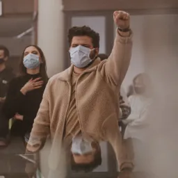 Mężczyzna w beżowej kurtce i maseczce podnosi zaciśniętą pięść podczas wydarzenia z innymi osobami w tle, wszyscy w maseczkach.
