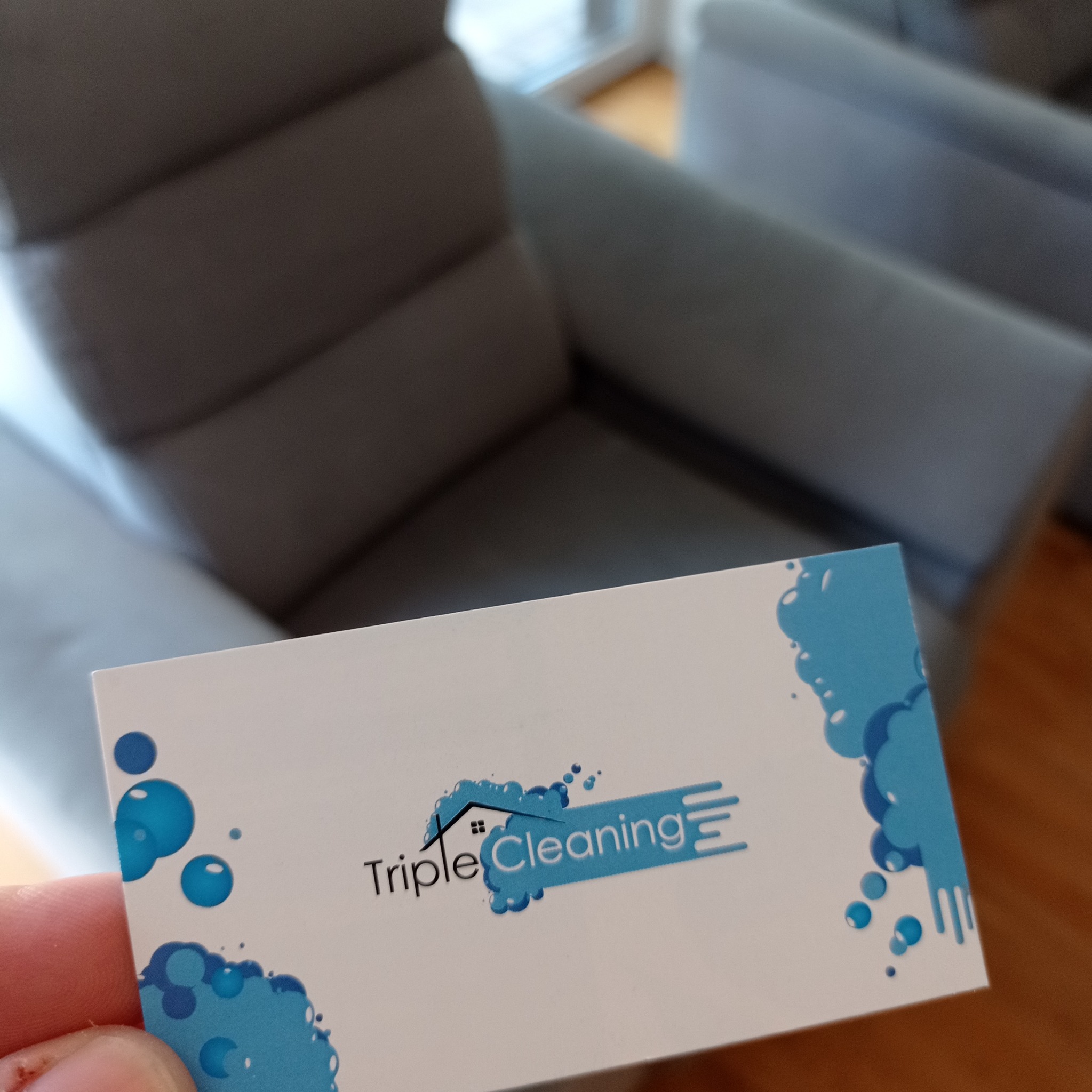 Wizytówka firmy sprzątającej 'Triple Cleaning' z logo w postaci domu otoczonego bąbelkami, umieszczona na tle szarego fotela.