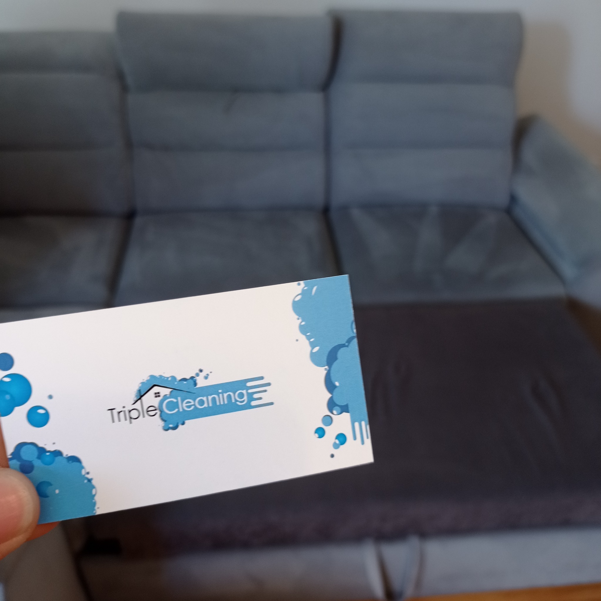 Wizytówka firmy Triple Cleaning na tle szarej, rozłożonej sofy.
