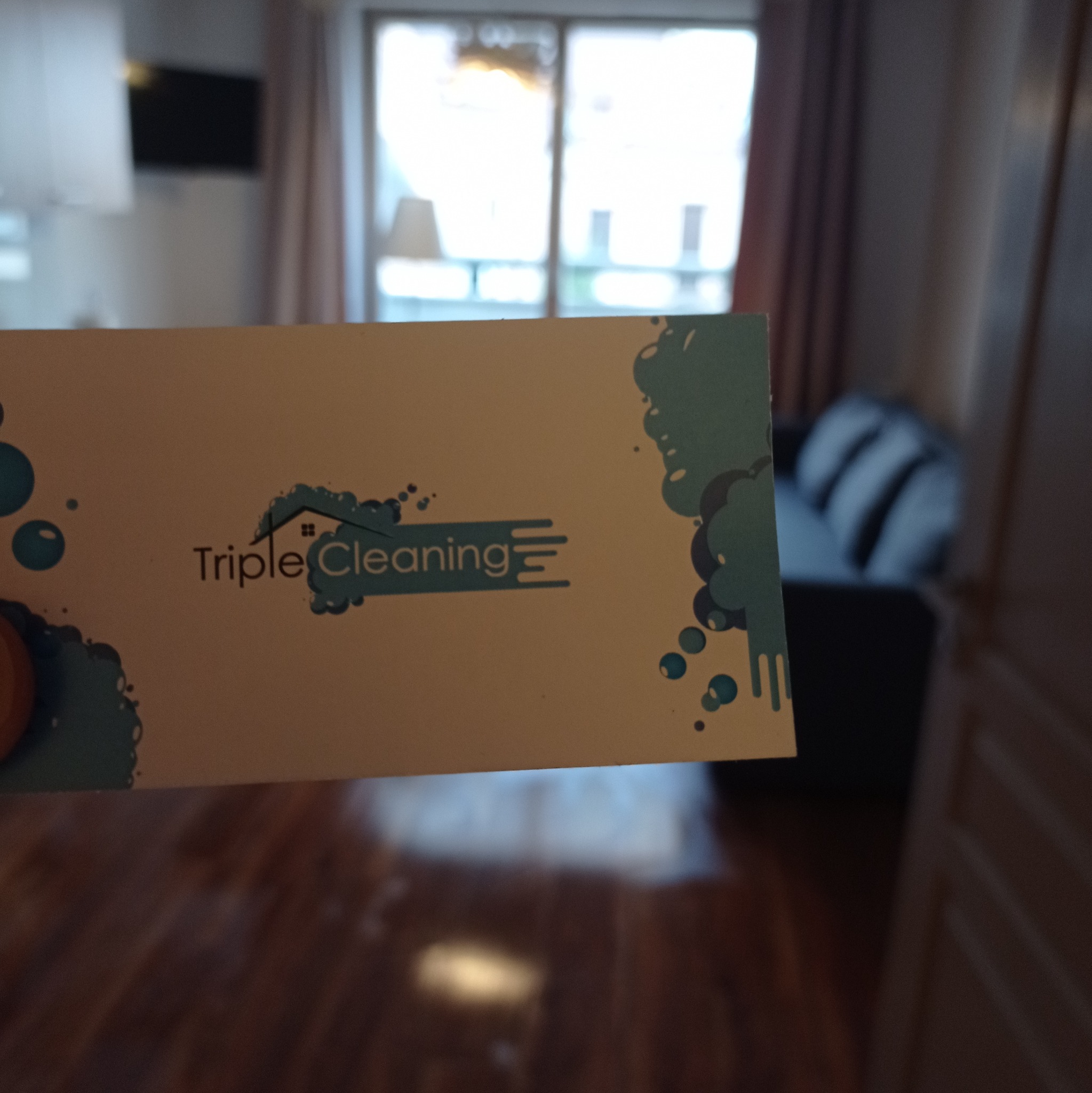 Wizytówka firmy sprzątającej 'Triple Cleaning' na tle wyczyszczonego mieszkania z drewnianą podłogą i szarą kanapą.
