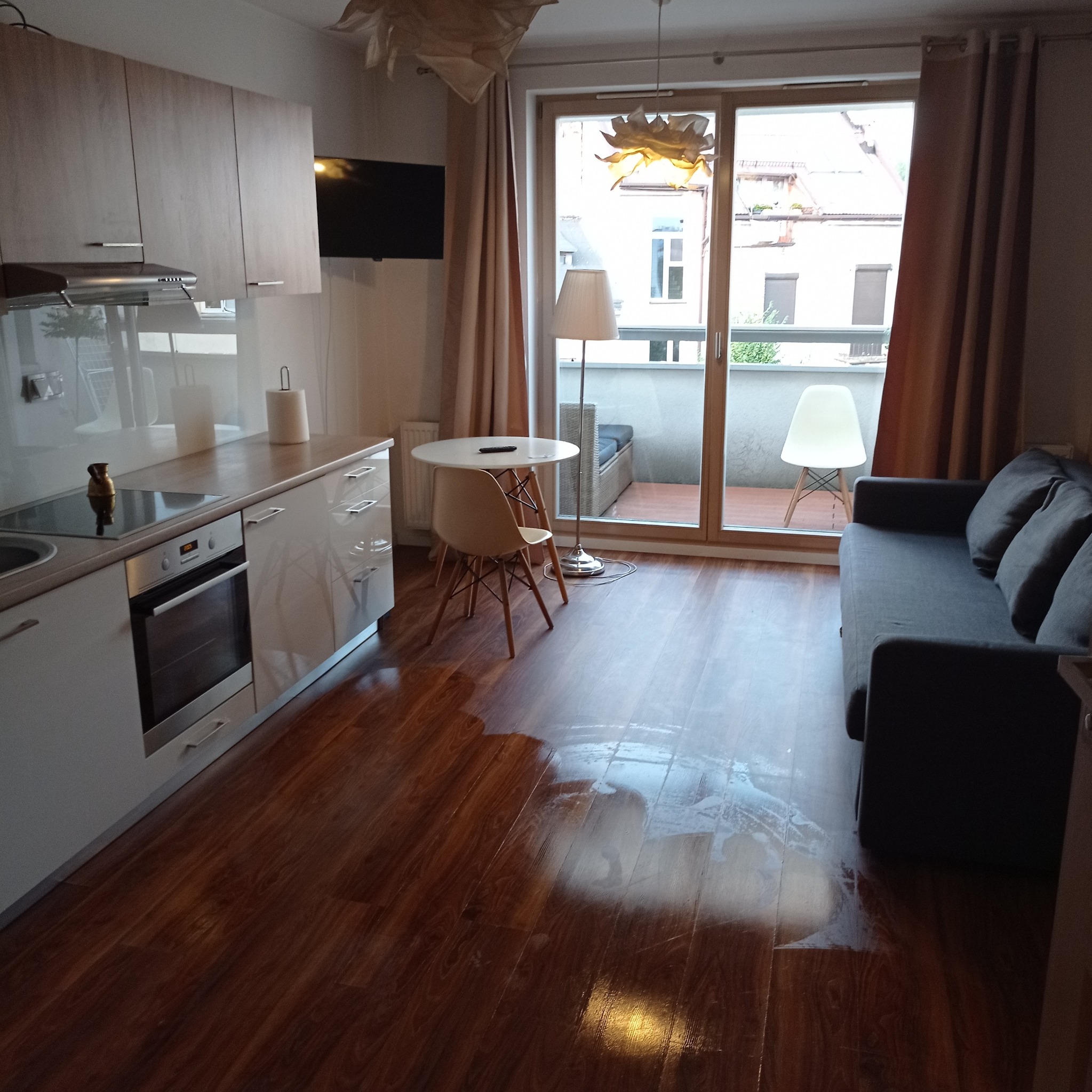 Wnętrze mieszkania w Krakowie: widok na salon z aneksem kuchennym i wyjściem na balkon; na podłodze widoczne ślady po umyciu, sugerujące świeżo zakończone prace porządkowe.