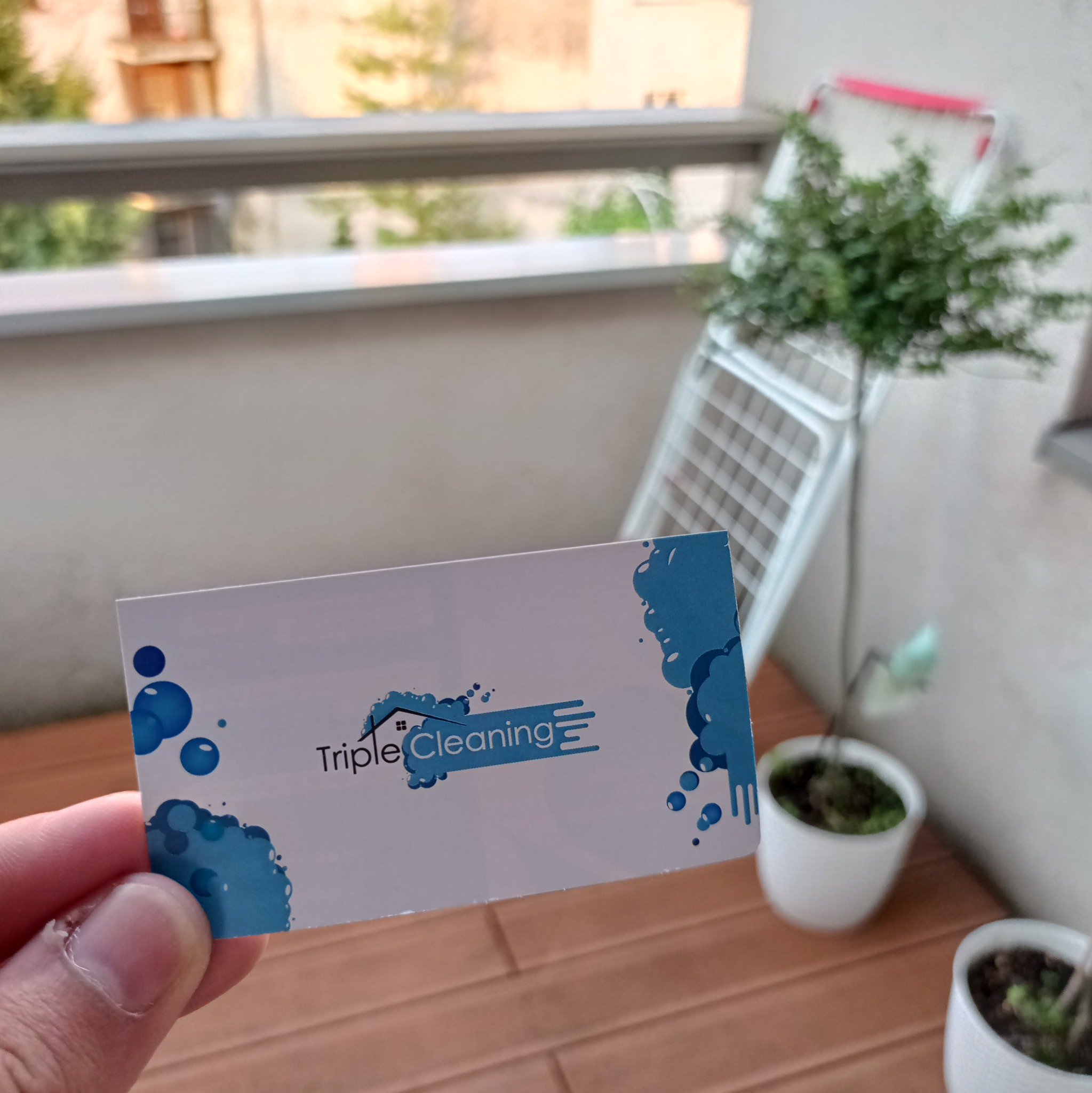 Wizytówka firmy sprzątającej Triple Cleaning z błękitnymi elementami graficznymi, trzymana w dłoni na tle balkonu z suszarką i roślinami w doniczkach.