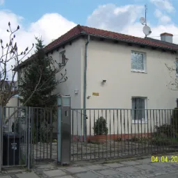 Dwukondygnacyjny dom z jasną elewacją i czerwoną dachówką, widoczny zza metalowego ogrodzenia, z anteną satelitarną na dachu i tabliczką z adresem Wilenska 71.