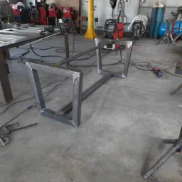 Metalowy szkielet stołu spawalniczego z kwadratowymi nogami w warsztacie, widoczne spawarki, butle z gazem i inne narzędzia.