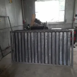 Metalowy panel ogrodzeniowy z pionowymi prętami, spawany w warsztacie, widoczne narzędzia i maszyny w tle, oświetlenie dzienne.