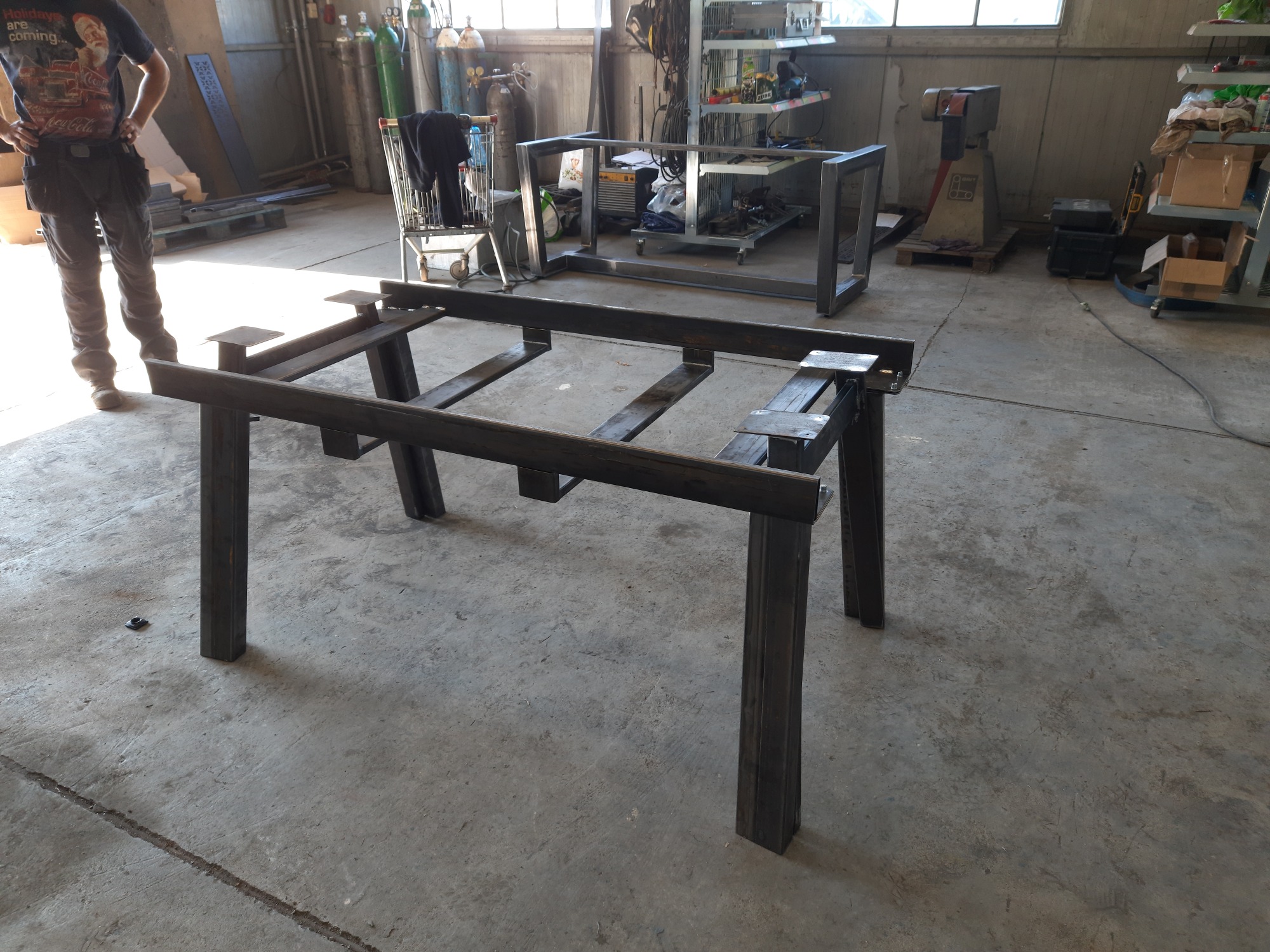 Stół z metalowych profili w warsztacie, widoczne elementy konstrukcyjne, spawarka, w tle wózek sklepowy i regał z narzędziami.