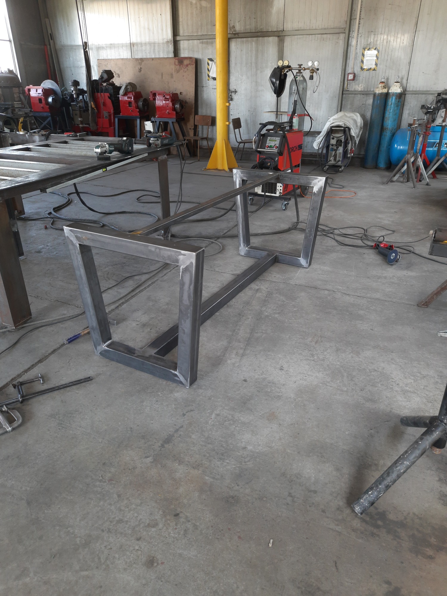 Metalowy szkielet stołu spawalniczego z kwadratowymi nogami w warsztacie, widoczne spawarki, butle z gazem i inne narzędzia.