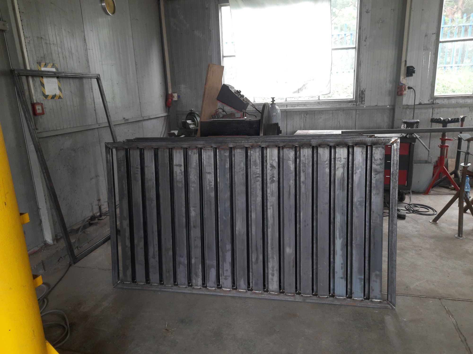 Metalowy panel ogrodzeniowy z pionowymi prętami, spawany w warsztacie, widoczne narzędzia i maszyny w tle, oświetlenie dzienne.