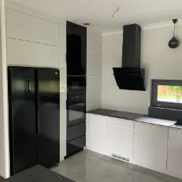 Nowoczesna kuchnia w bieli i czerni z dużym, dwudrzwiowym lodówko-zamrażarką, zabudowanymi piekarnikami i płytą grzewczą, okapem oraz oknem z widokiem na zieleń.