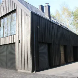 Nowoczesny dom z ciemną, pionową elewacją drewnianą, metalowym dachem i szarymi roletami zewnętrznymi, otoczony drzewami.