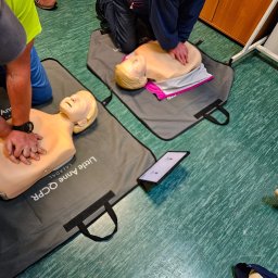 PROMEDYK - MACIEJ DĘBICKI - Instruktorzy demonstrujący resuscytację krążeniowo-oddechową na manekinach Little Anne QCPR podczas kursu pierwszej pomocy. Widoczny tablet z instrukcjami.