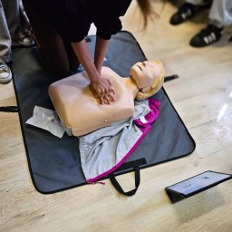 PROMEDYK - MACIEJ DĘBICKI - Kurs pierwszej pomocy w Kalwarii Zebrzydowskiej: ćwiczenia resuscytacji na manekinie z widocznym tabletem z instrukcjami i uczestnikami szkolenia w tle.