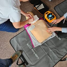 PROMEDYK - MACIEJ DĘBICKI - Kurs pierwszej pomocy: Instruktorzy ćwiczą resuscytację na manekinie Little Anne QCPR z użyciem defibrylatora AED Trainer. Widoczne notatki i akcesoria.