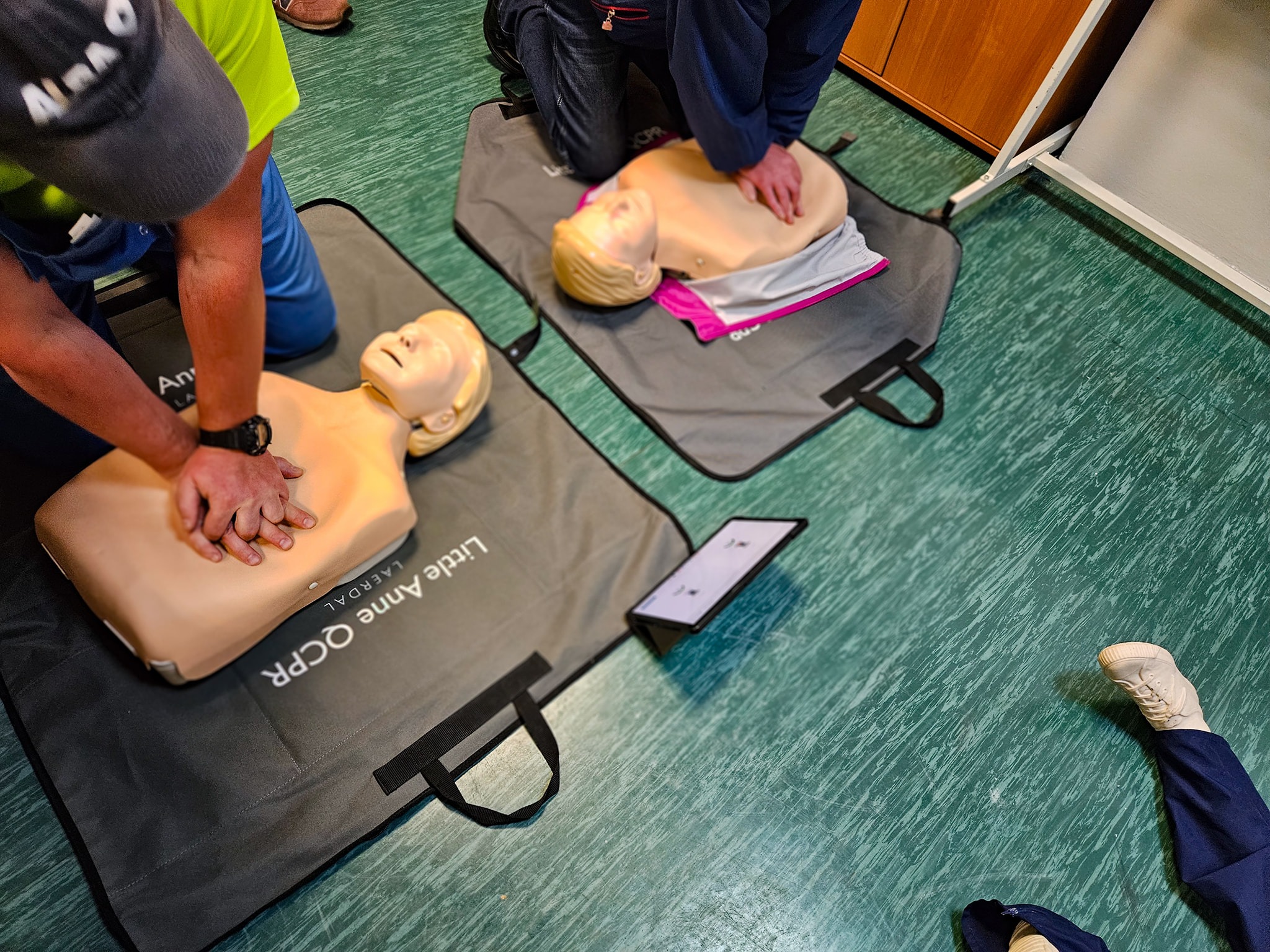 Instruktorzy demonstrujący resuscytację krążeniowo-oddechową na manekinach Little Anne QCPR podczas kursu pierwszej pomocy. Widoczny tablet z instrukcjami.