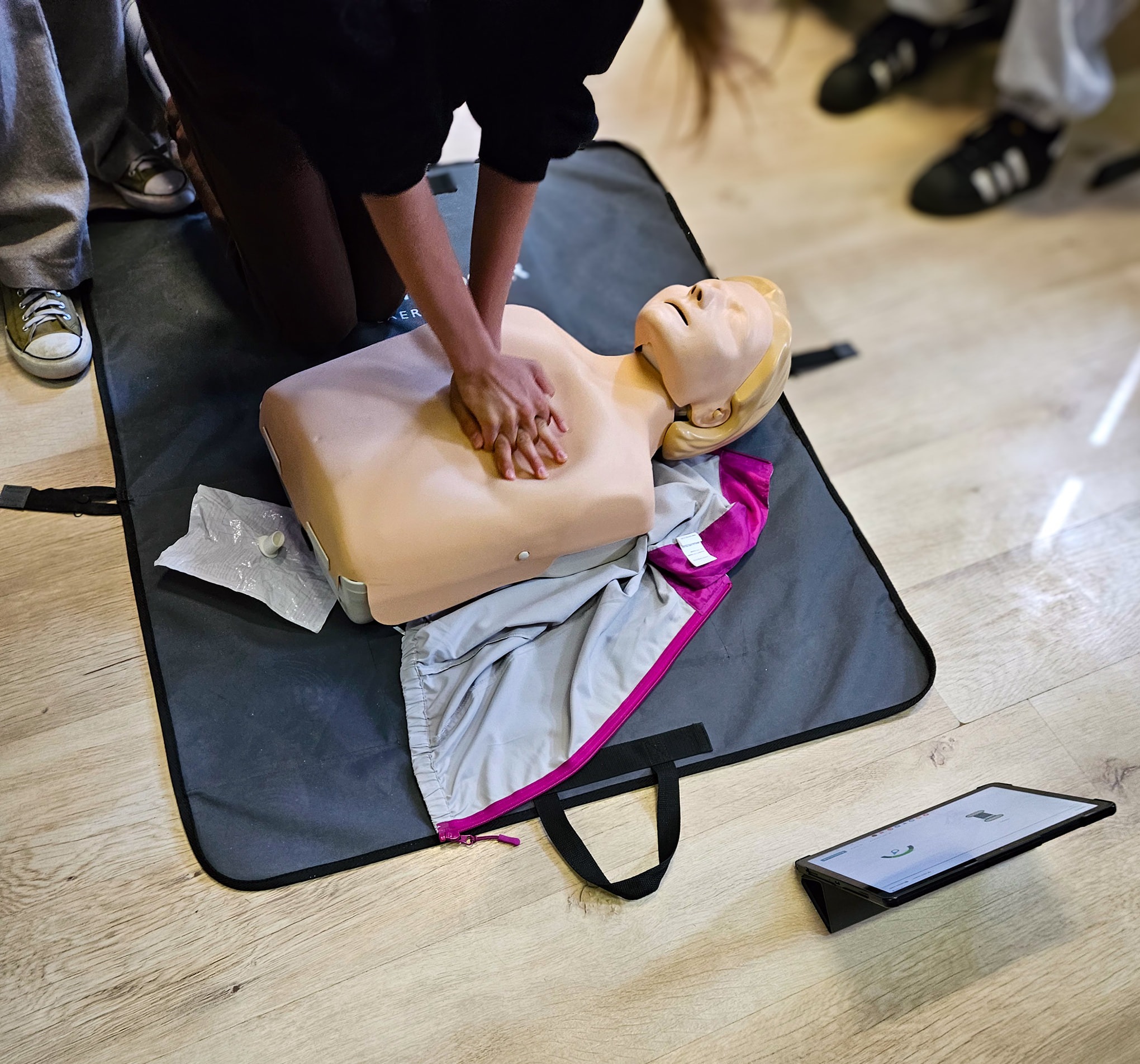 Kurs pierwszej pomocy w Kalwarii Zebrzydowskiej: ćwiczenia resuscytacji na manekinie z widocznym tabletem z instrukcjami i uczestnikami szkolenia w tle.